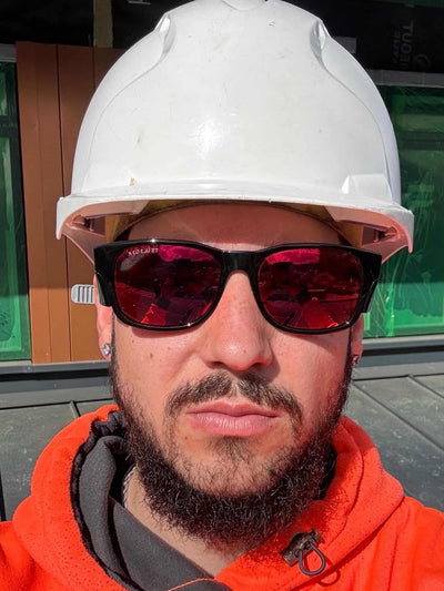 Tradesperson in white hard hat and orange hi-vis wearing Trade Shades black frame safety glasses with red polarised lenses on site